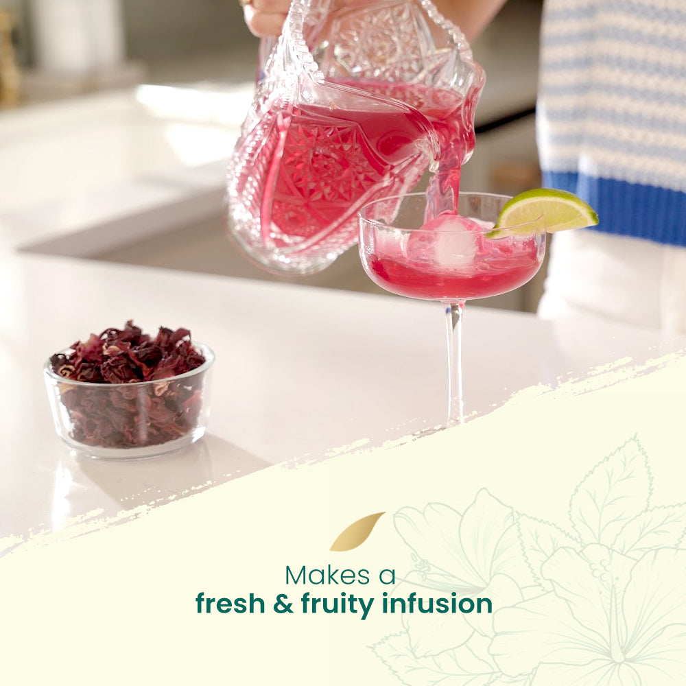 Pink hibiscus cocktail being poured into a glass with a lime wedge and dried fruits on a kitchen counter.